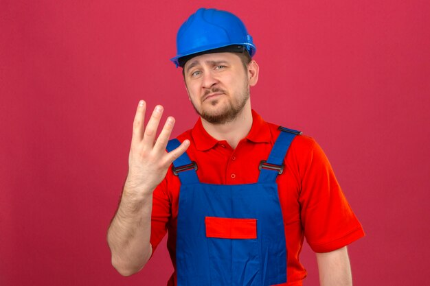 Человек-строитель в строительной форме и защитном шлеме показывает и указывает пальцами номер четыре со скептическим выражением лица над изолированной розовой стеной