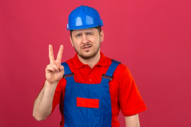 Builder man wearing construction uniform and security helmet showing and pointing up with fingers number two with skeptic expression on face over isolated pink wall