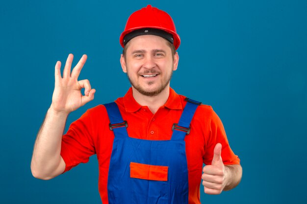 Builder man wearing construction uniform and security helmet smiling friendly doing ok sign and showing thumb up over isolated blue wall