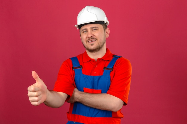 Человек-строитель в строительной форме и защитном шлеме дружелюбно улыбается, показывая большой палец вверх, стоя над изолированной розовой стеной