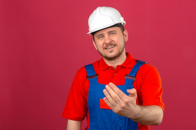 Foto gratuita uniforme da costruzione dell'uomo del costruttore e casco di sicurezza che sorridono facendo un gesto con la mano che invita a venire sopra la parete rosa isolata