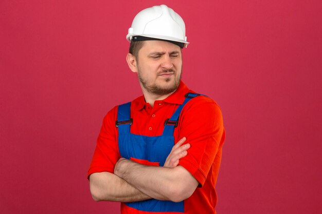 Мужчина-строитель в строительной форме и каске безопасности стоит со скрещенными руками и смотрит в сторону с хмурым лицом, демонстрируя неприязнь к изолированной розовой стене