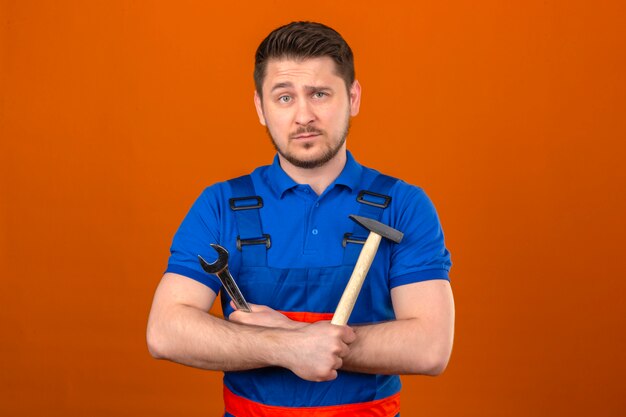 Builder man wearing construction uniform and security helmet standing with arms crossed with hammer and wrench in hands with serious face over isolated orange wall