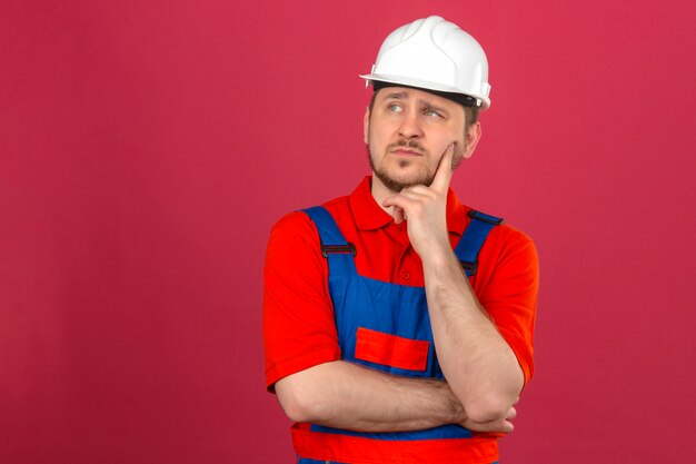 작성기 남자 건설 유니폼과 보안 헬멧을 착용 그의 뺨을 만지고 생각에 잠겨있는 모습 격리 된 분홍색 벽에 서