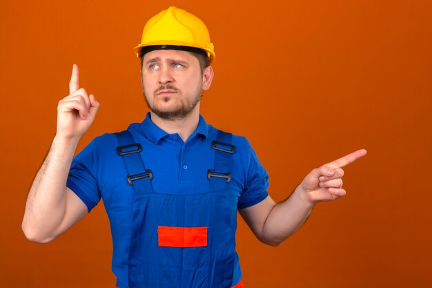 Builder man wearing construction uniform and security helmet warns of danger lifting his finger up showing warning sign with finger standing over isolated orange wall