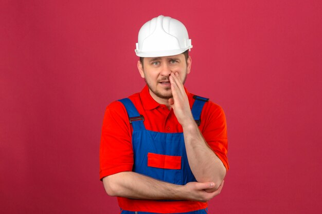 고립 된 어두운 분홍색 벽 위에 비밀 서 말하는 입 근처 손으로 건설 유니폼 및 보안 헬멧을 착용 작성기 남자