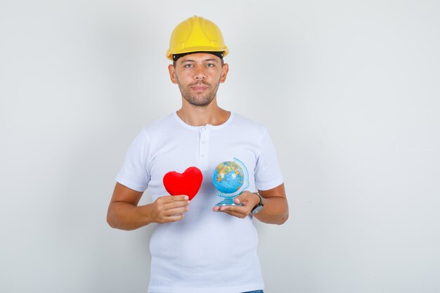 Builder man in white t-shirt, helmet holding red heart and globe, front view.