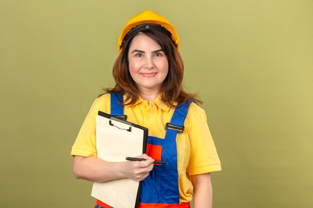 Free photo builder woman in construction uniform and safety helmet holding clipboard and pen looking confident with smile on face over isolated green wall