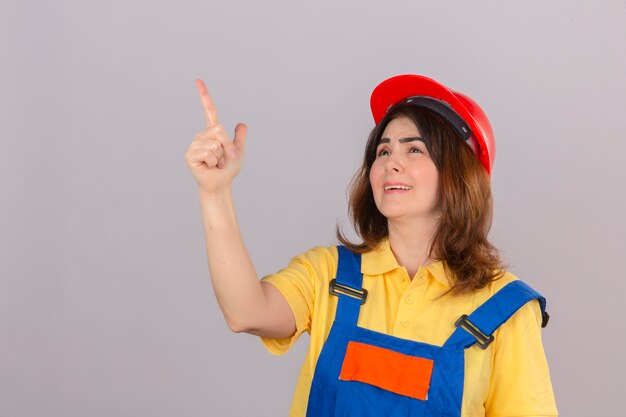 Женщина-строитель в строительной форме и защитном шлеме смотрит вверх, указывая пальцем в сторону, улыбаясь над изолированной белой стеной