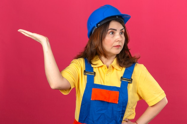 Builder woman in construction uniform and safety helmet shrugging shoulders raising hand not understanding what happened clueless and confused expression over isolated pink wall