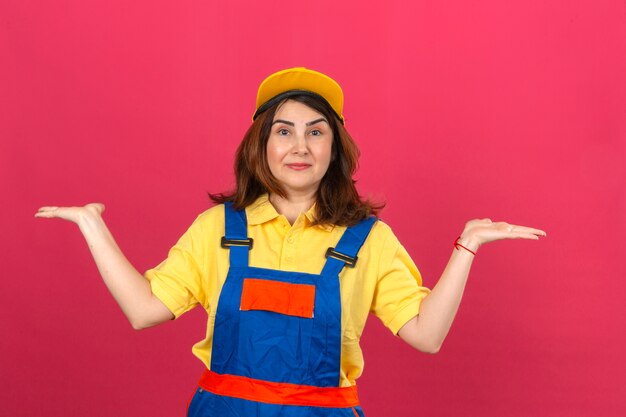 Builder woman in construction uniform and safety helmet shrugging shoulders spreading hands not understanding what happened clueless and confused expression over isolated pink wall