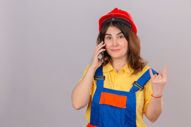 Женщина-строитель в строительной форме и защитном шлеме разговаривает по мобильному телефону, делая рок-символ пальцами над изолированной белой стеной