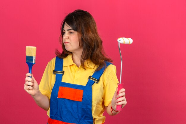 Builder woman wearing construction uniform holding paint roller and brush looking at it with sad expression displeased over isolated pink wall