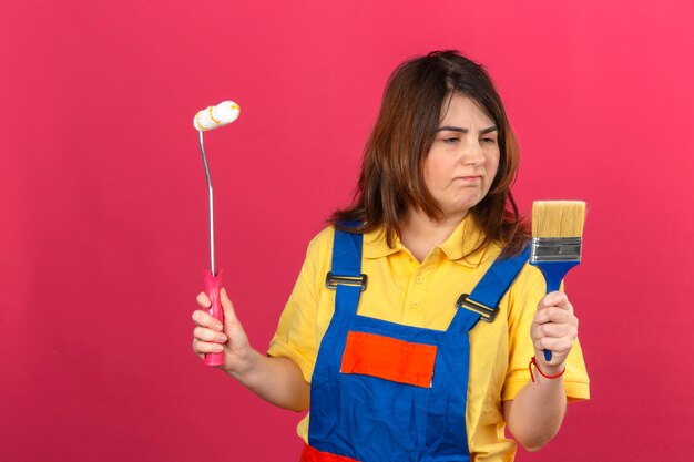 Rullo e spazzola di pittura d'uso della tenuta dell'uniforme della costruzione della donna del costruttore che la esaminano con l'espressione triste dispiaciuta sopra la parete rosa isolata