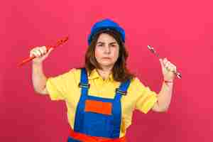 Free photo builder woman wearing construction uniform and safety helmet holding adjustable wrenches in raised hands with angry expression over isolated pink wall