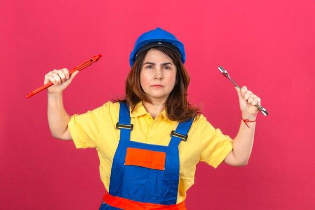 Builder woman wearing construction uniform and safety helmet holding adjustable wrenches in raised hands with angry expression over isolated pink wall