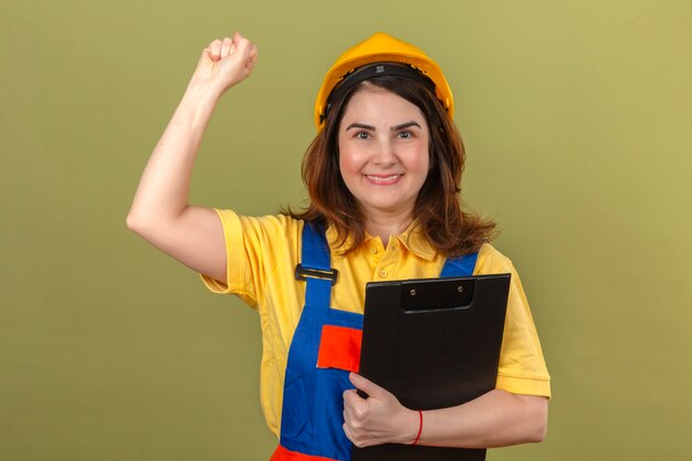 Builder woman wearing construction uniform and safety helmet holding clipboard with smile raising fist after a victory winner concept over isolated green wall