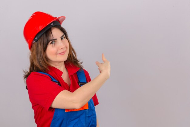 Builder woman wearing construction uniform and safety helmet indicate something at blank copy space behind her over isolated white wall