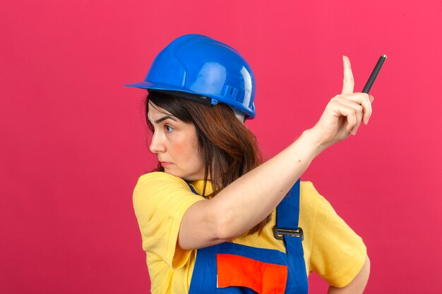 Builder woman wearing construction uniform and safety helmet looking asides pointing with finger up warning over isolated pink wall
