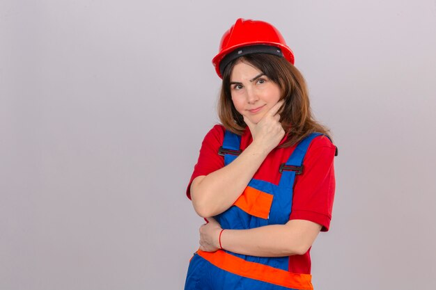 Builder woman wearing construction uniform and safety helmet looking confident at the camera smiling standing with crossed arms and hand raised on chin thinking positive over isolated white backgr