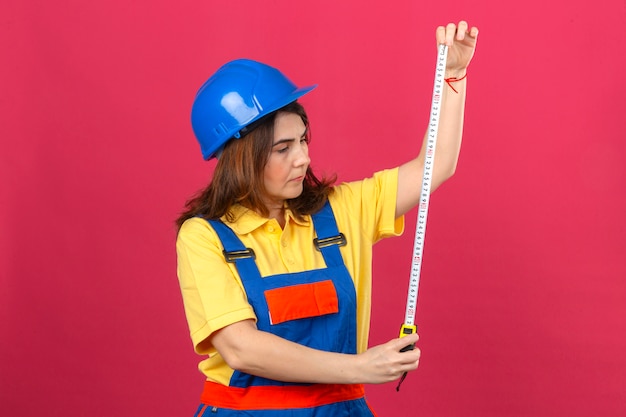 Builder woman wearing construction uniform and safety helmet looking at measurement tape in hands with serious face over isolated pink wall