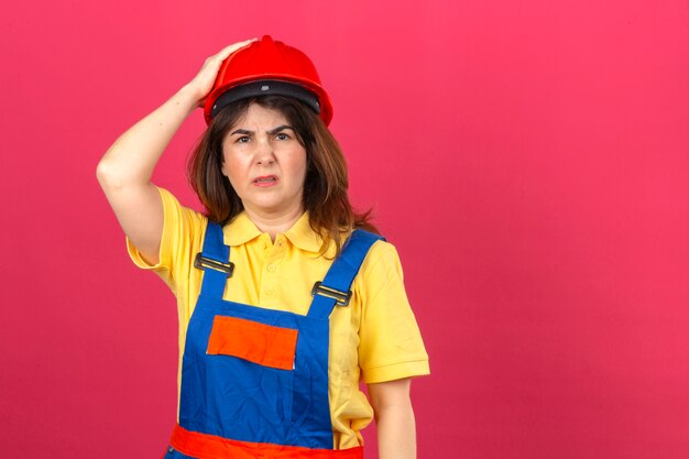 Builder woman wearing construction uniform and safety helmet looking unwell with sad expression touching head having headache over isolated pink wall