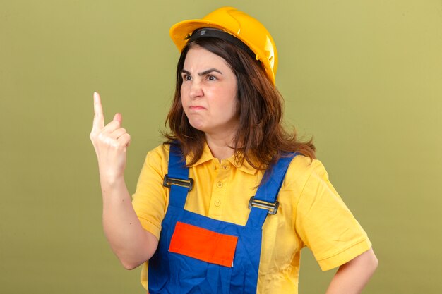 Builder woman wearing construction uniform and safety helmet pointing finger up looking reproachfully at someone over isolated green wall