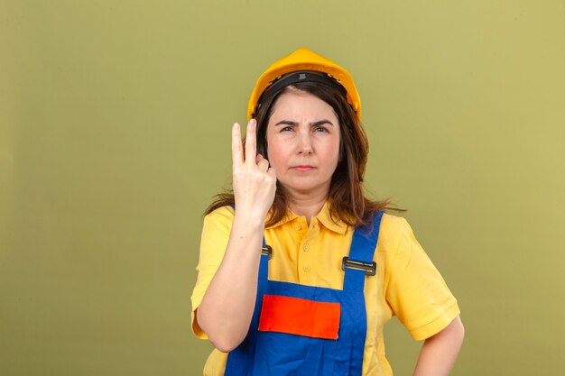 Builder woman wearing construction uniform and safety helmet showing and pointing up fingers number two with suspicious expression standing over isolated green wall