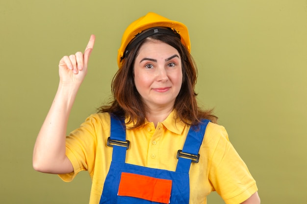 Builder woman wearing construction uniform and safety helmet showing and pointing up with finger while smiling confident and happy standing over isolated green wall