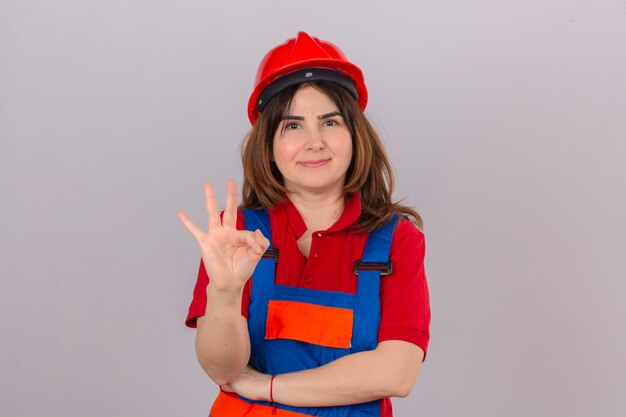 Builder woman wearing construction uniform and safety helmet smiling friendly showing and pointing up with fingers number three over isolated white wall