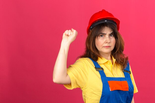 Женщина-строитель в строительной форме и защитном шлеме с сердитым выражением лица поднимает кулак над изолированной розовой стеной