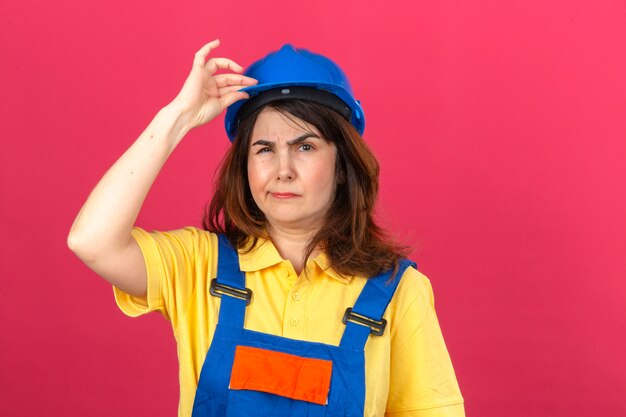 Женщина-строитель в строительной форме и защитном шлеме со скептическим выражением лица трогает шлем, стоящий над изолированной розовой стеной