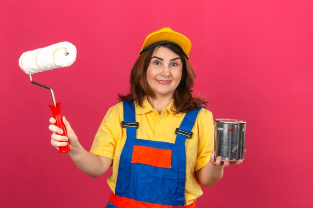 Женщина-строитель в строительной форме и желтой кепке держит малярный валик и банку с краской, улыбаясь со счастливым лицом над изолированной розовой стеной