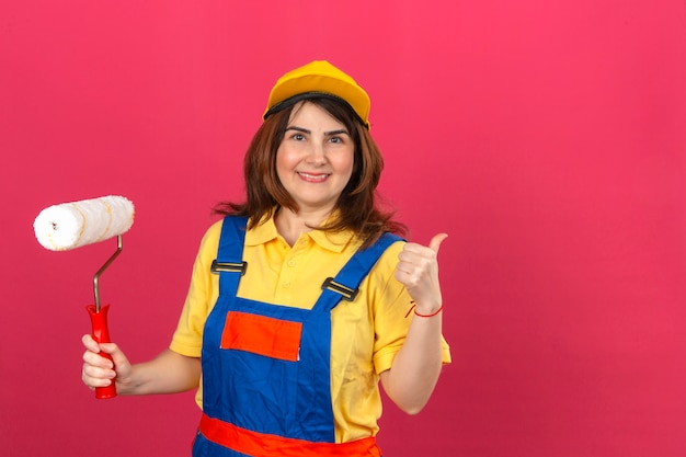 Uniforme edile d'uso della costruzione della donna del costruttore e rullo di pittura della tenuta dello spiritello malevolo e mostrare pollice su sorridere allegro sopra la parete rosa isolata