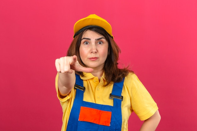 Женщина-строитель в строительной форме и желтой кепке выглядит удивленно, указывая пальцем на камеру над изолированной розовой стеной