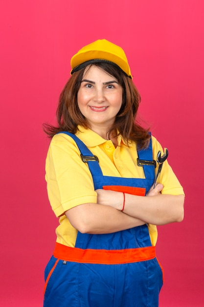 작성기 여자 건설 유니폼과 무기와 노란 모자 서 입고 고립 된 분홍색 벽에 쾌활 한 미소 렌치로 넘어