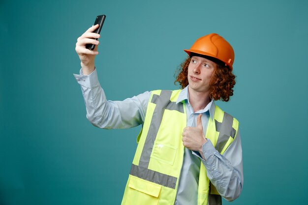 건설 유니폼을 입은 빌더 청년과 파란색 배경 위에 서 있는 엄지손가락을 보여주는 스마트폰 미소를 사용하여 셀카를 하는 안전 헬멧