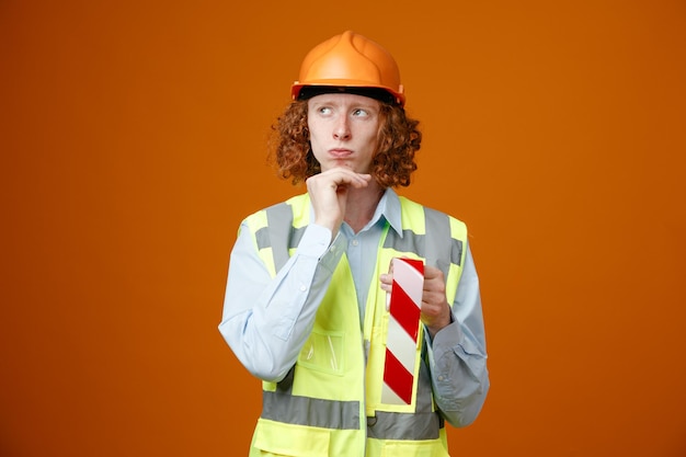 Молодой человек-строитель в строительной форме и защитном шлеме держит липкую ленту, смотрит в сторону с задумчивым выражением лица и думает, положив руку на подбородок, стоя на оранжевом фоне