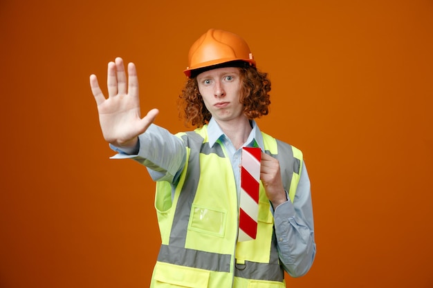Builder young man in construction uniform and safety helmet holding adhesive tape looking at camera worried making stop gesture with hand standing over orange background