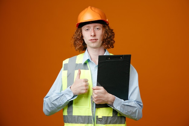 Builder young man in construction uniform and safety helmet holding clipboard looking at camera smiling confident showing thumb up standing over orange background