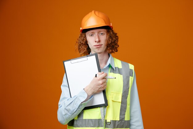 Giovane costruttore in uniforme da costruzione e casco di sicurezza che tiene appunti e pennarello guardando la fotocamera con espressione sicura in piedi su sfondo arancione