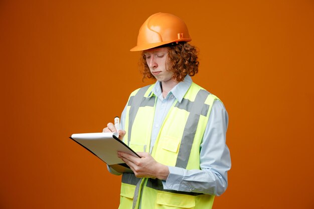 Builder young man in construction uniform and safety helmet holding clipboard and marker writing something looking confident standing over orange background