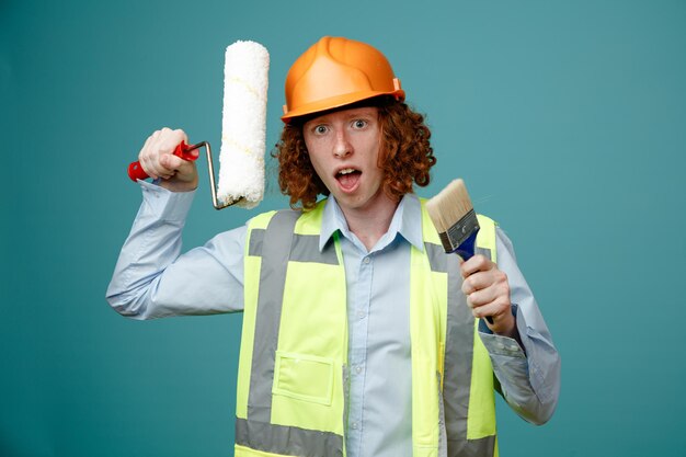 Builder young man in construction uniform and safety helmet holding paint roller and brush looking at camera being surprised standing over blue background
