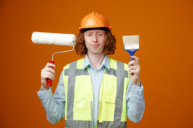 Builder young man in construction uniform and safety helmet holding paint roller and brush looking at camera smiling confident standing over orange background
