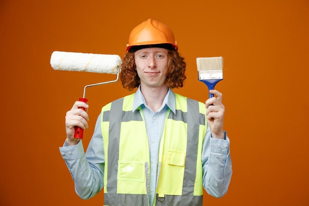 Builder young man in construction uniform and safety helmet holding paint roller and brush looking at camera smiling confident standing over orange background