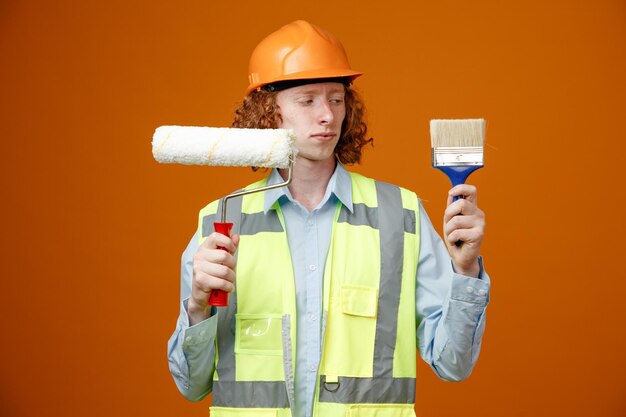 Builder young man in construction uniform and safety helmet holding paint roller and brush looking confused trying to make choice standing over orange background