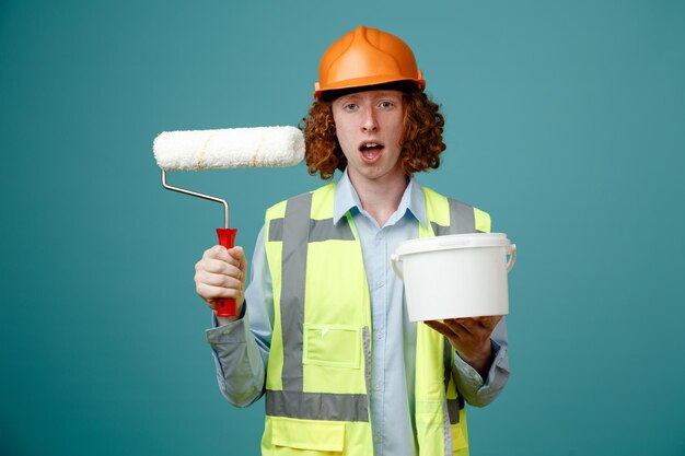 Builder young man in construction uniform and safety helmet holding paint roller and bucket looking surprised standing over blue background