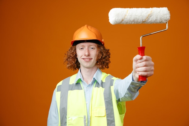 Молодой человек-строитель в строительной форме и защитном шлеме держит валик с краской и смотрит в камеру, улыбаясь уверенно, счастливо и позитивно, стоя на оранжевом фоне