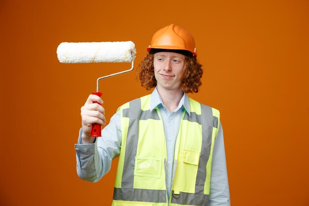 Builder young man in construction uniform and safety helmet holding paint roller looking at it happy and pleased smiling standing over orange background