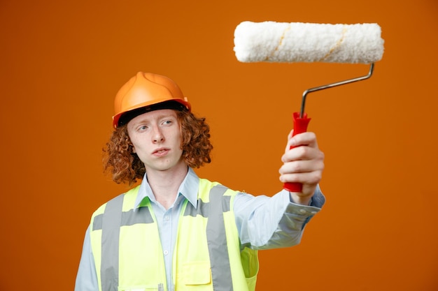 Builder young man in construction uniform and safety helmet holding paint roller looking at it with pensive expression standing over orange background